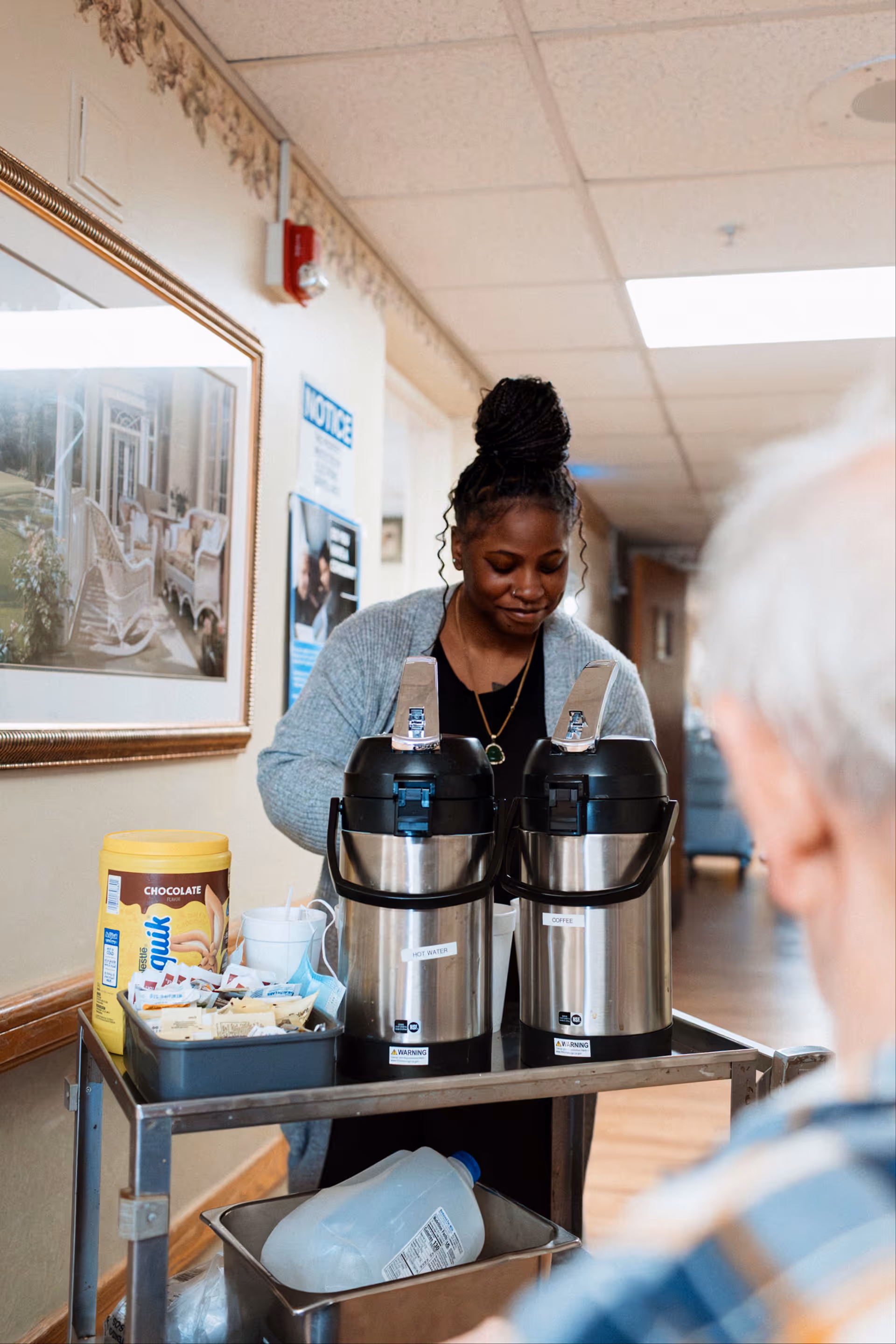 Caregiver prepares drinks on a service cart in a senior living facility hallway.