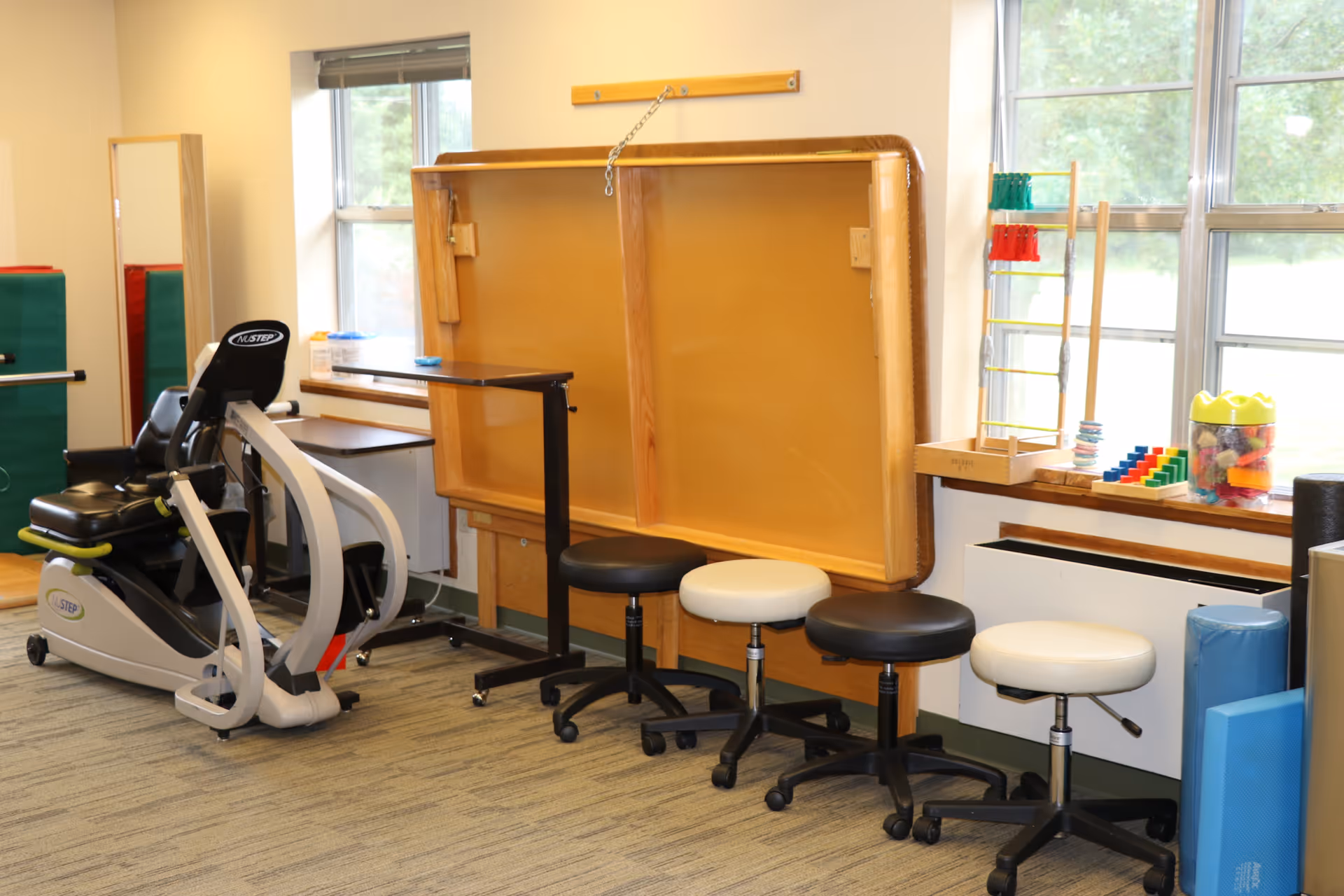 Indoor senior facility therapy room with exercise bike, stools, windows, and a large padded board.