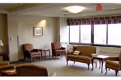 A bright senior living lounge with sofas, chairs, coffee tables, and large windows.