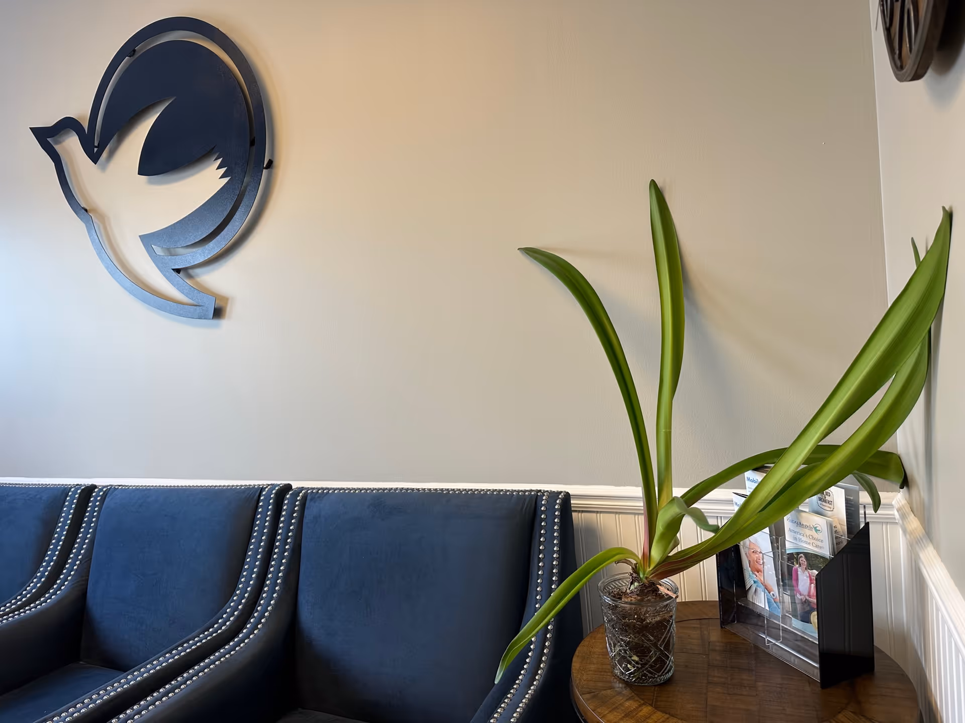 Blue upholstered sofa with nailhead trim, potted plant, and brochures beside a circular wall logo.