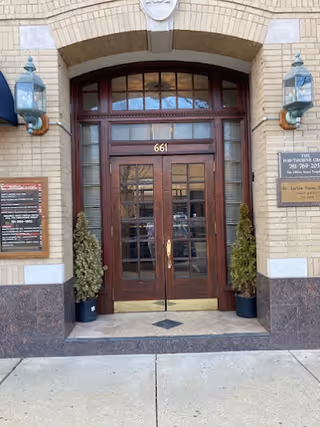 Double wooden doors framed by brick facade, arched transom, flanked by lanterns and potted shrubs.