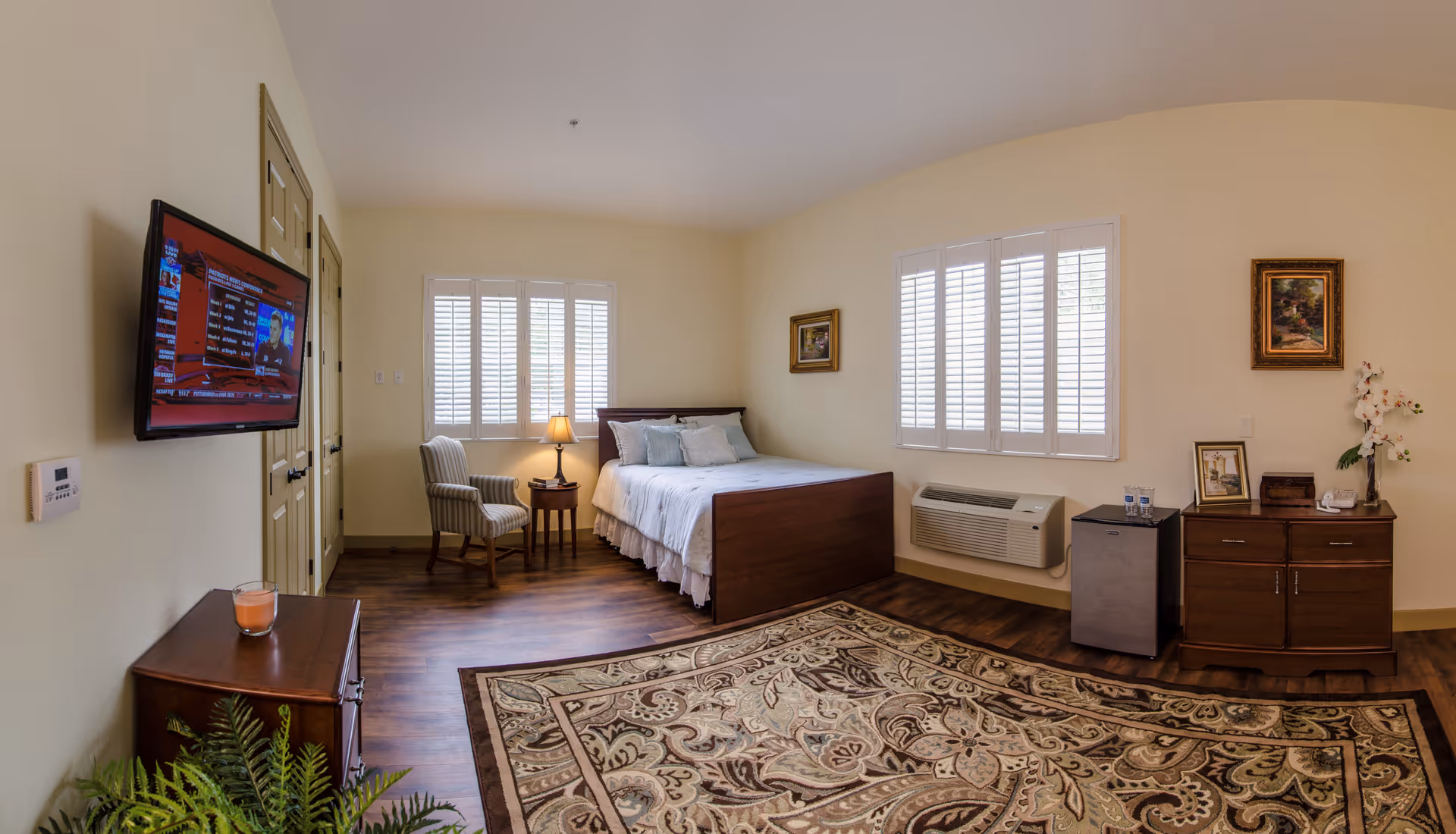 A senior living facility bedroom with a wooden bed, white bedding, two windows with white shutters, a small round table with a lamp and an armchair, a wall-mounted TV, a wooden dresser with a candle, a mini fridge, and a wooden cabinet with decorative items and framed pictures. The room has hardwood floors and a large patterned area rug.