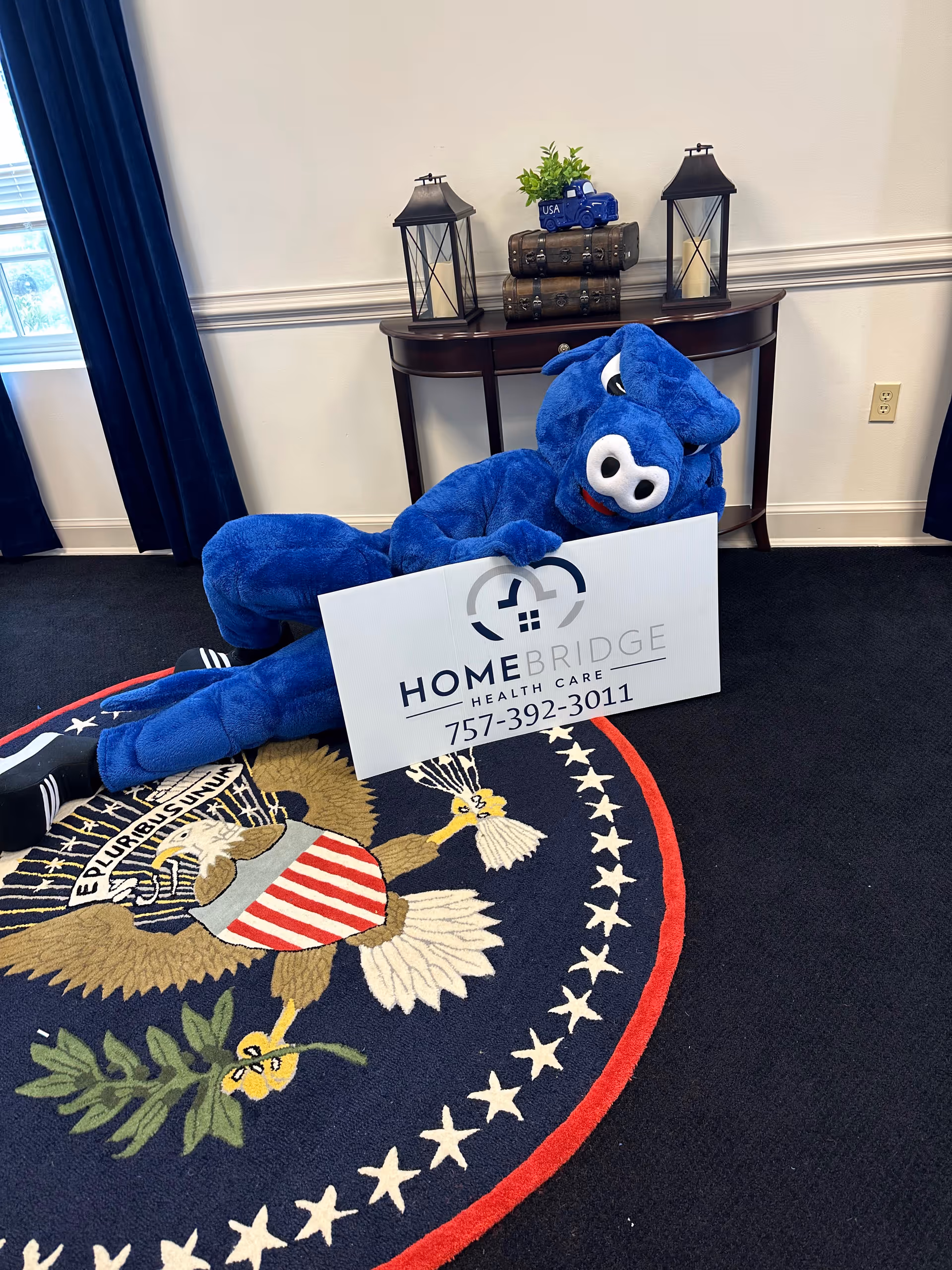Blue plush mascot holding a Homebridge Health Care sign inside a lobby.