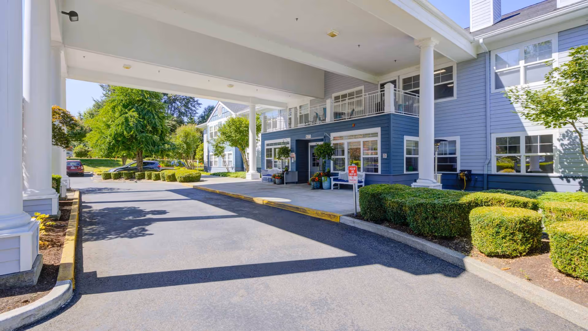 Entrance area of The Sequoia Assisted Living Community showing a covered driveway with white columns, blue building exterior, neatly trimmed bushes, and parked cars in the background under a clear sky.
