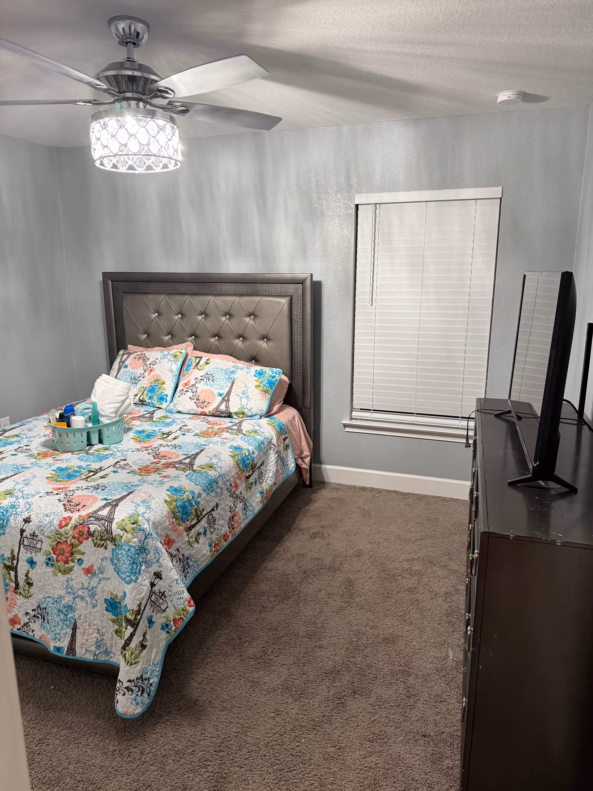 Small bedroom with a patterned bed, tufted headboard, dresser, window blinds, and ceiling fan.