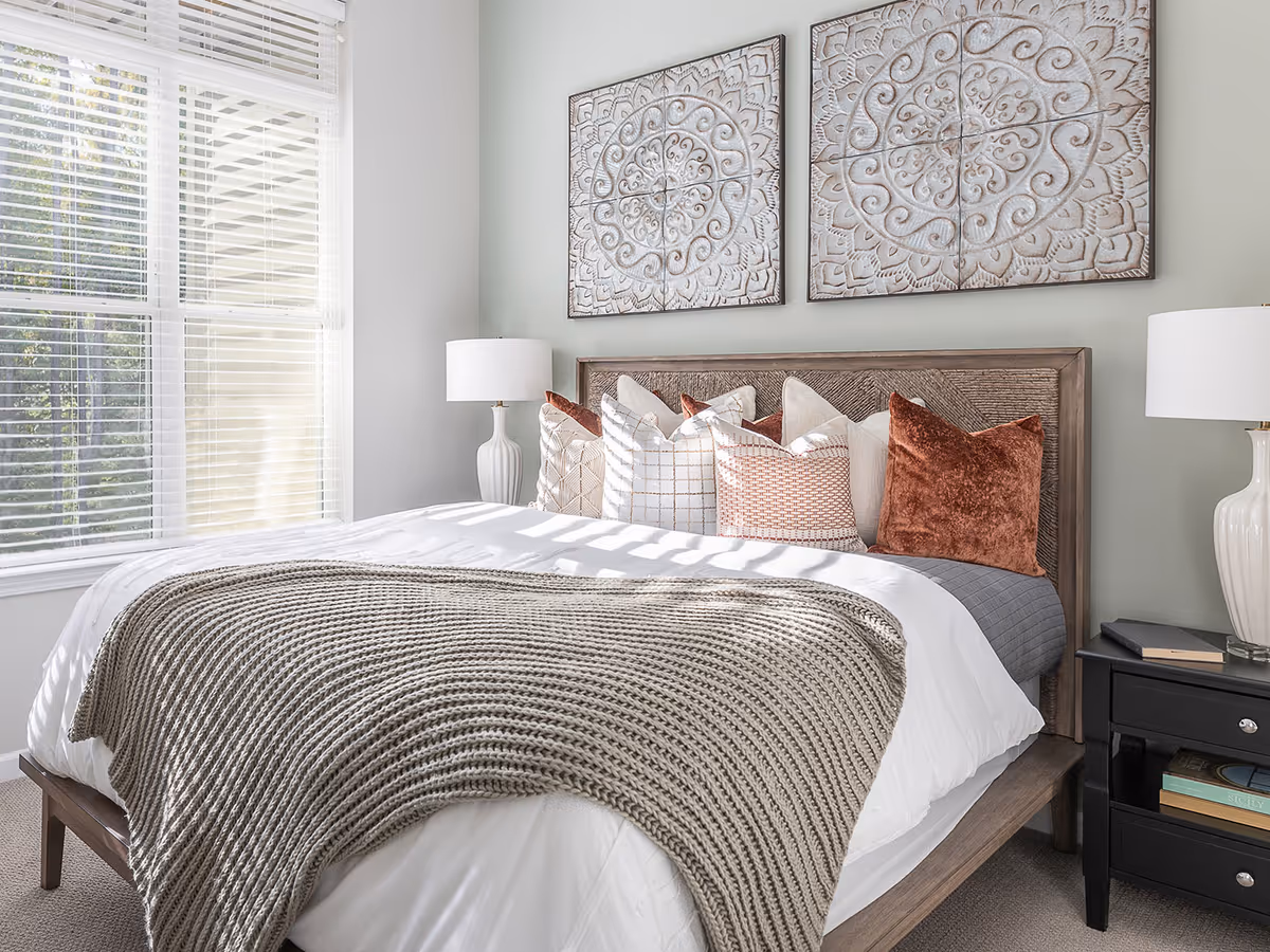A neatly made bedroom with a wooden bed, pillows, lamps, and large window blinds.