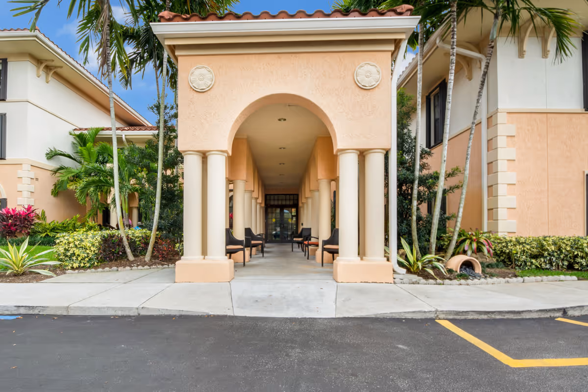 Entrance of a senior living facility with a covered walkway supported by columns, outdoor chairs lined along the sides, surrounded by tropical plants and palm trees, with peach-colored walls and a tiled roof.