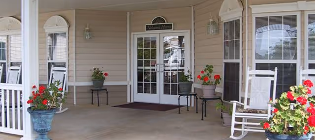 Front entrance of senior living facility with double doors, potted red flowers, and rocking chair.