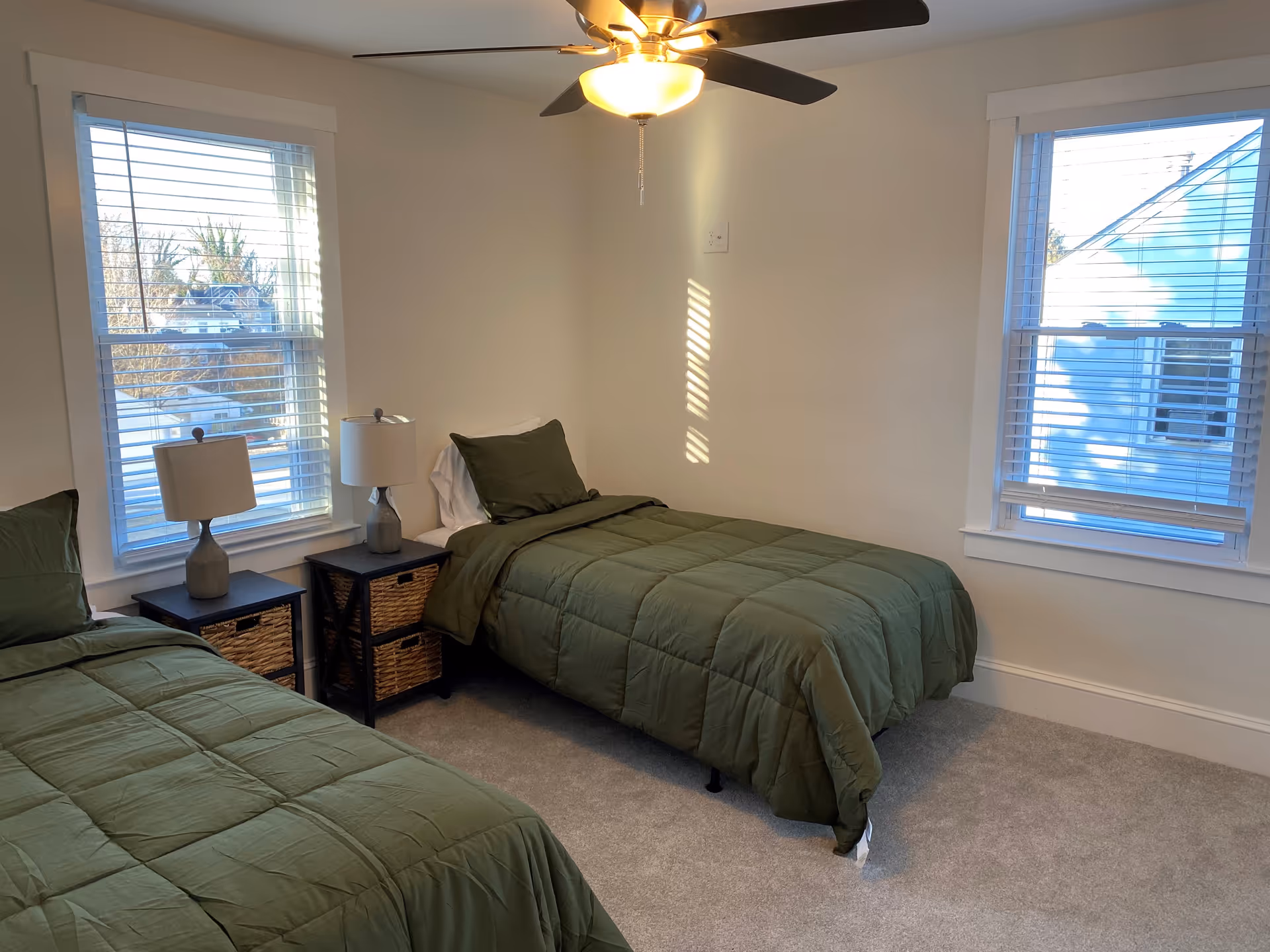 Two twin beds with green comforters, nightstands, lamps, and windows in a small bedroom.