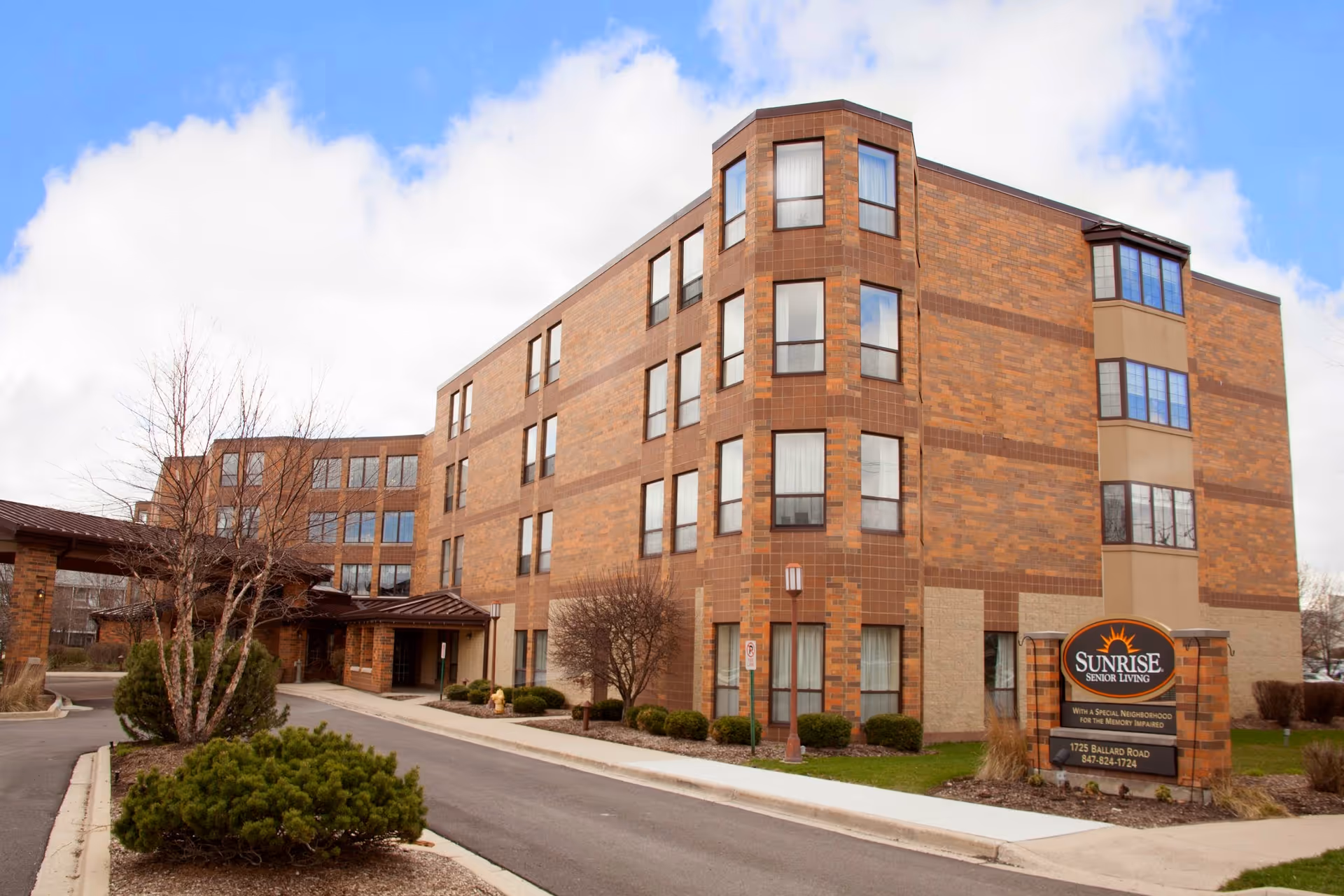 Exterior view of a multi-story brick building with multiple windows, a driveway, and landscaped bushes and trees. A sign in front reads 'Sunrise Senior Living' with additional information about a special neighborhood for the memory impaired.