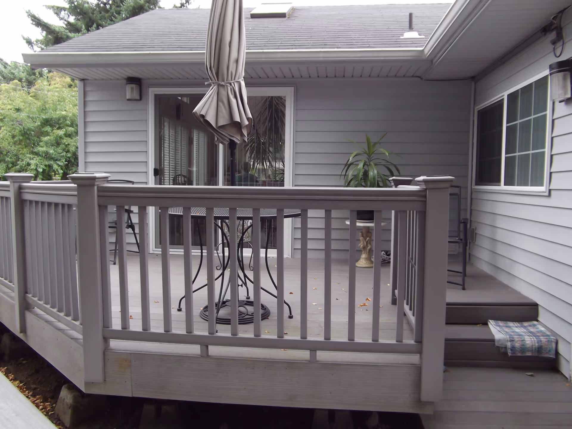 A covered outdoor patio area with gray wooden railings and flooring. There is a round metal table with a closed umbrella in the center, a potted plant on a pedestal, and a few chairs. The patio is attached to a gray house with sliding glass doors and windows.