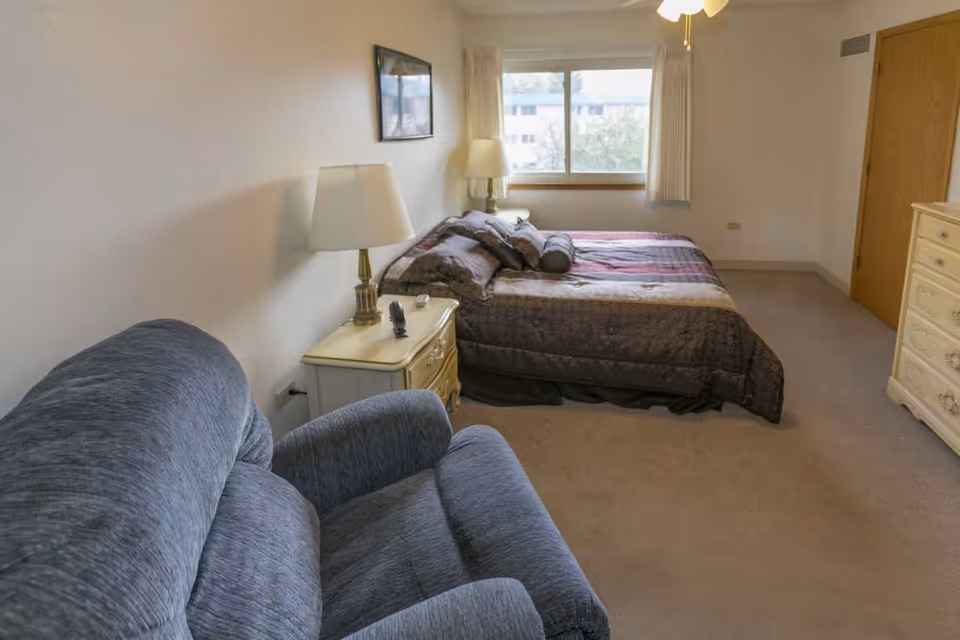 A sunlit bedroom featuring a bed with patterned bedding, a recliner, nightstands with lamps, and a dresser.