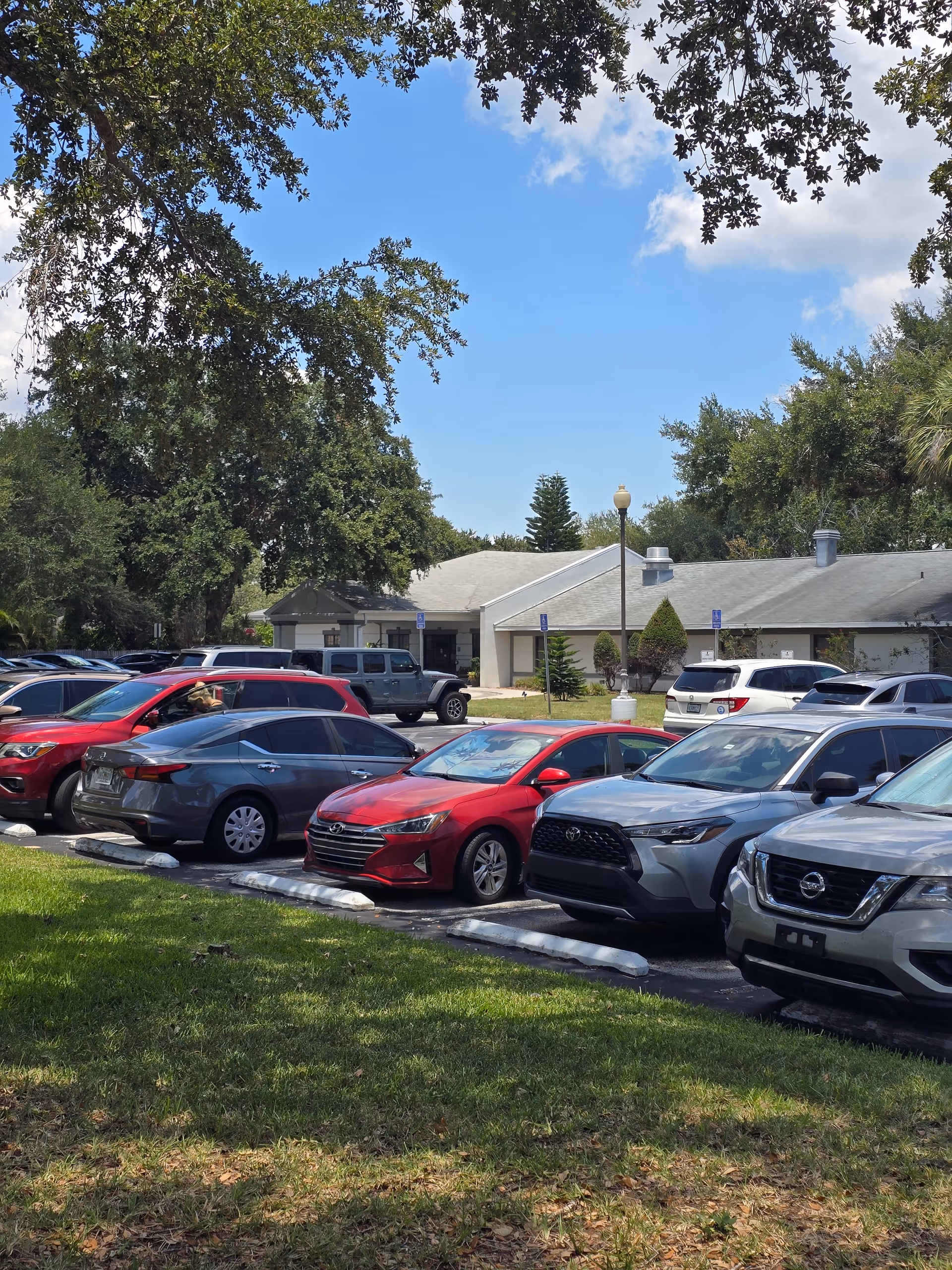 Parking lot in front of a single-story light building with trees and parked cars.