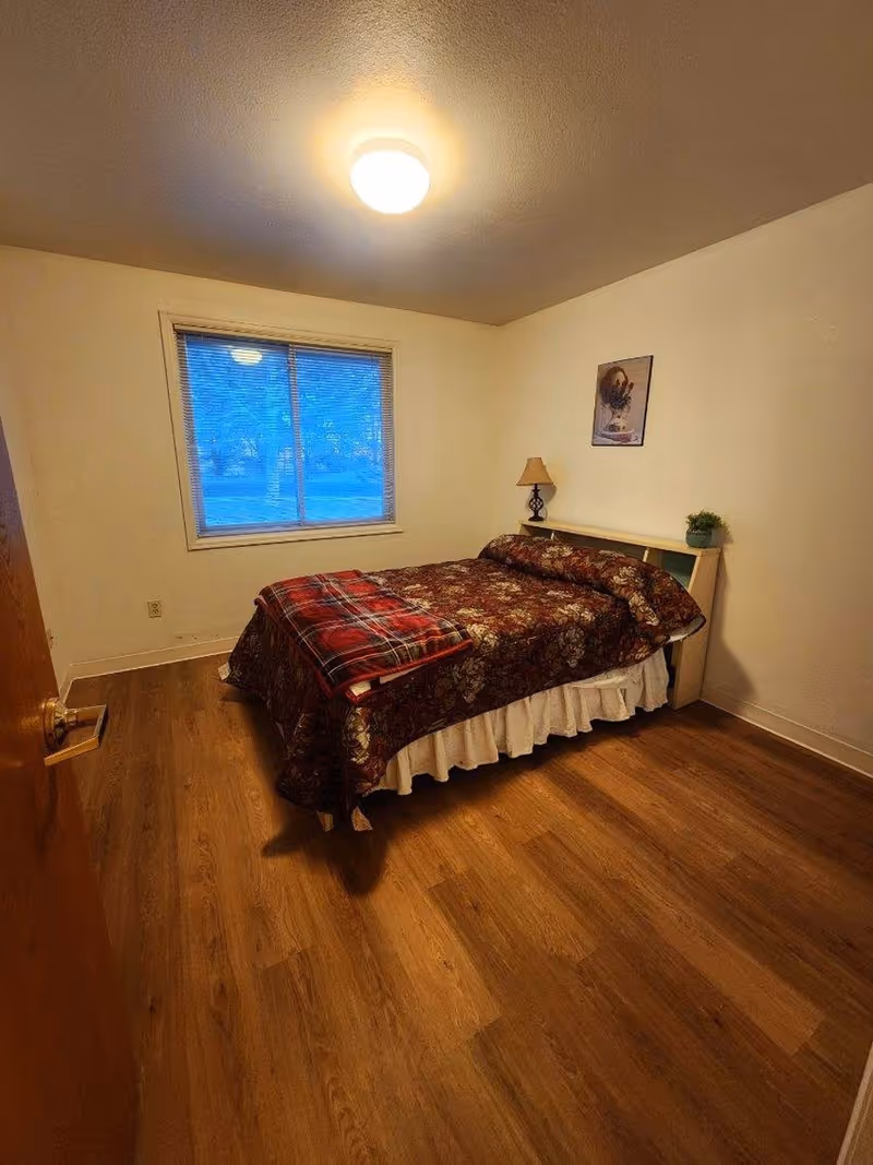 Cozy bedroom with a bed, nightstand lamp, window, and wood flooring.
