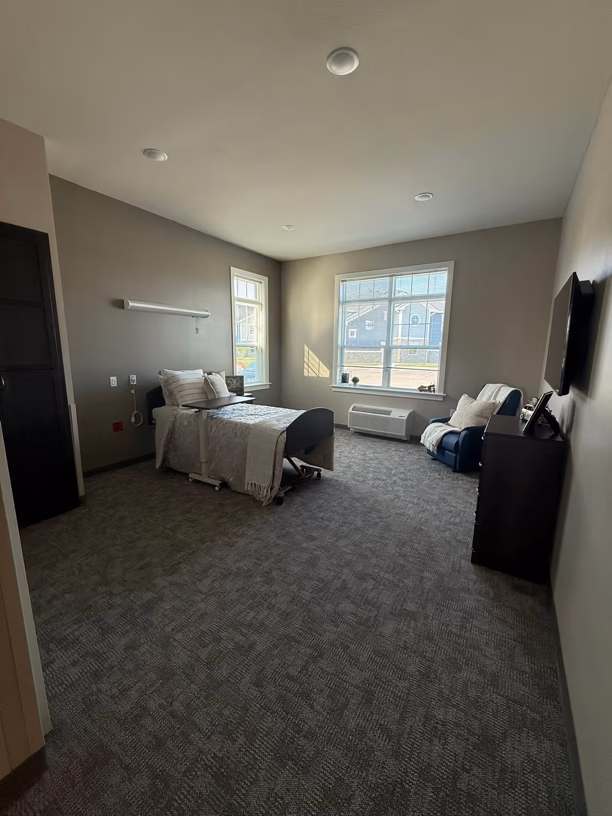 A modern senior living bedroom with a bed, chair, dresser, and large windows.
