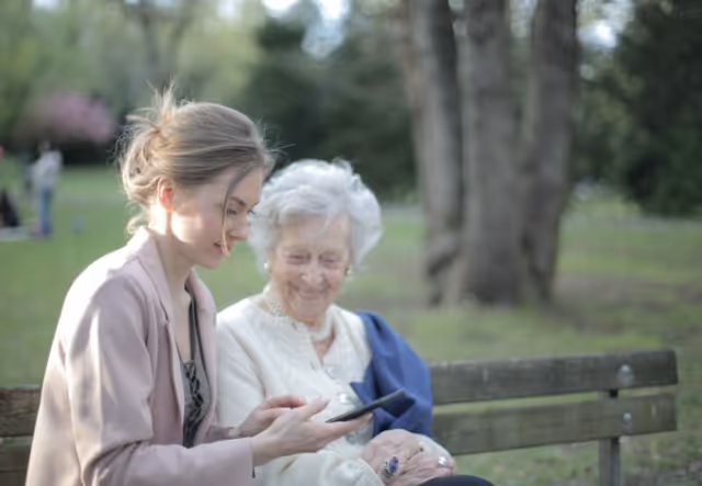 A caregiver and an elderly woman sit on a park bench outdoors, looking at a smartphone.