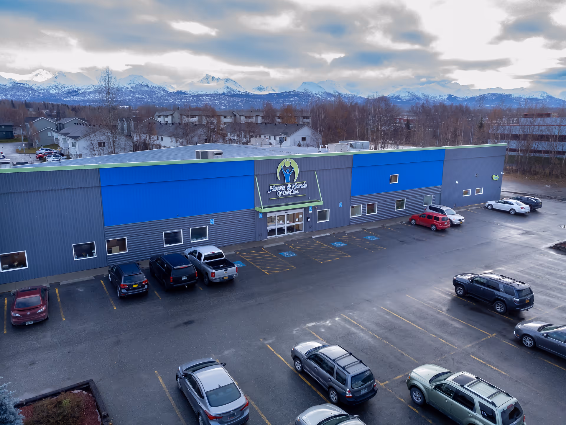 Aerial view of a blue-gray care facility with a parking lot and mountain backdrop