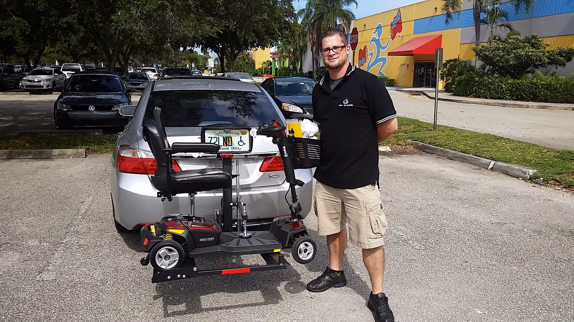Man with a mobility scooter attached to a car in a parking lot by a building.