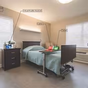 Hospital-style bed in a small furnished bedroom with a desk, laptop, and dresser.