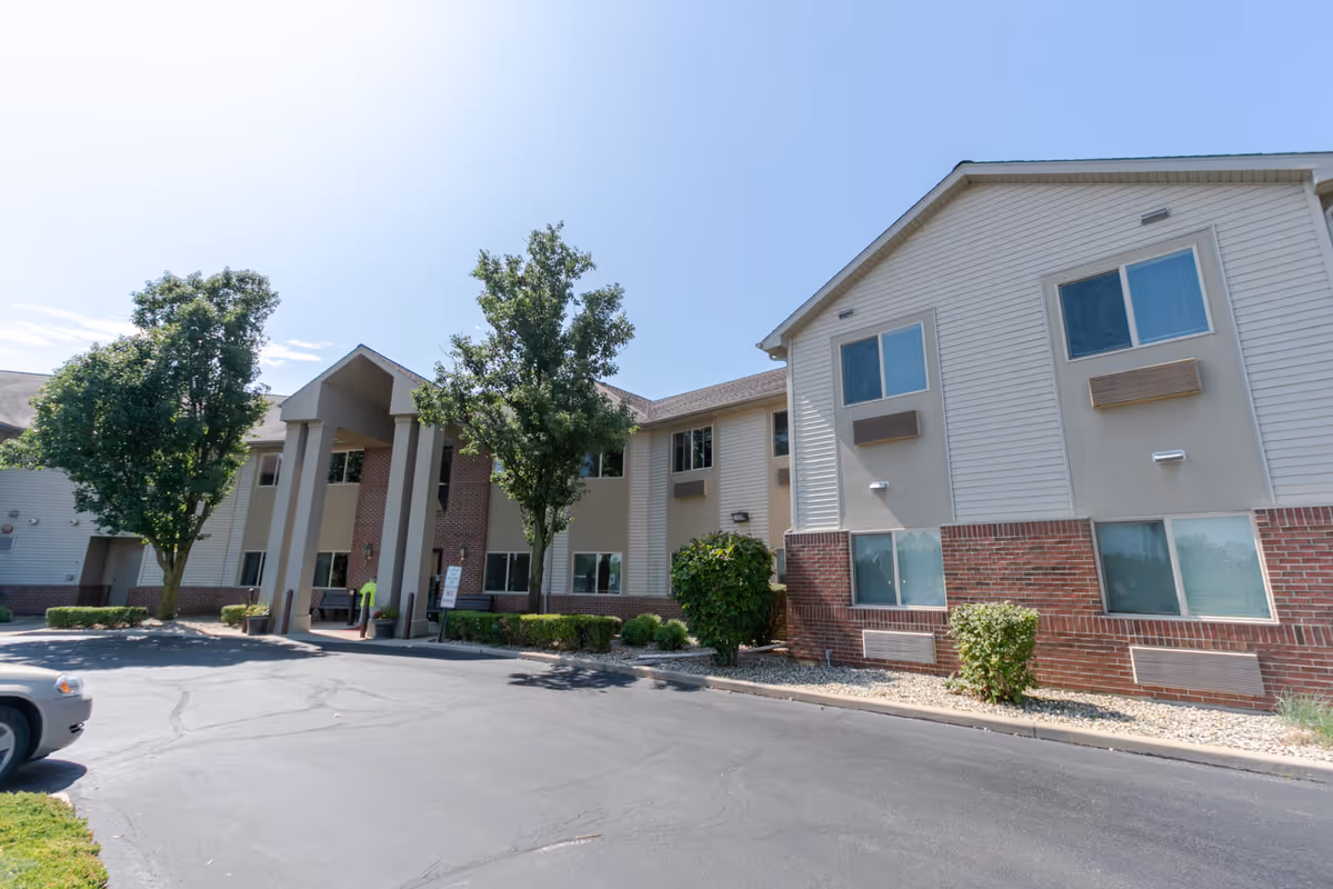 Exterior view of a two-story senior living facility building with beige siding and red brick accents. The entrance features a covered portico supported by four columns. There are several windows on both floors, trees, and shrubs around the building, and a paved driveway in front.