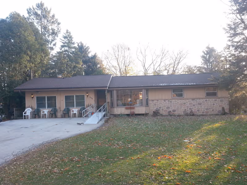 Front exterior of a single-story ranch-style building with a concrete driveway, a wheelchair ramp, porch chairs, and a grassy yard with trees.
