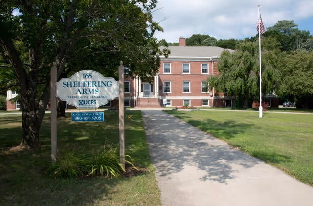 Walkway and front lawn leading to a two-story brick retirement residence with a sign that reads 'Sheltering Arms'.