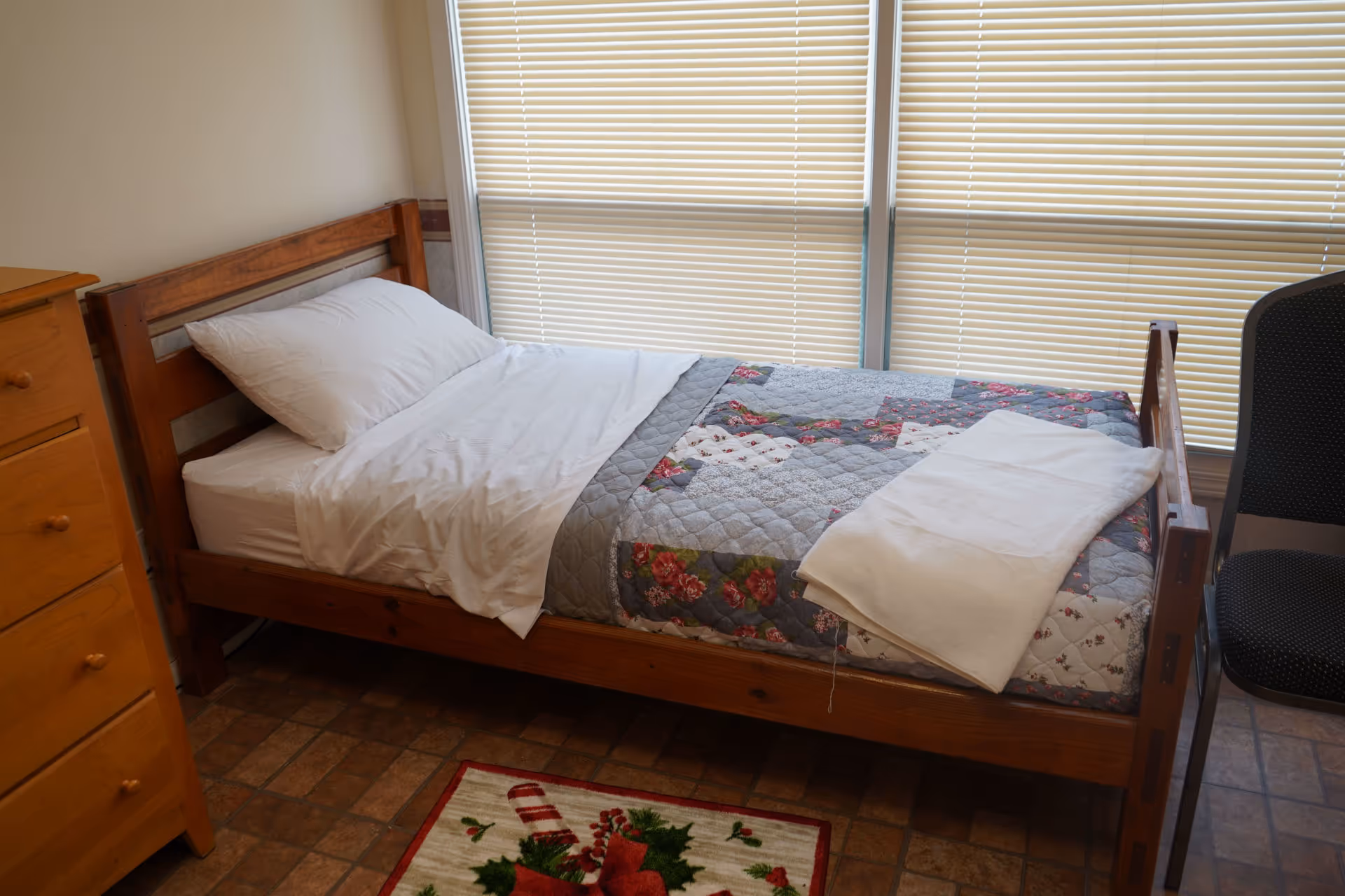 Wooden single bed with quilt, pillow, dresser, blinds, and a chair in a small bedroom.