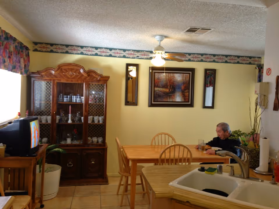 An elderly man sits at a wooden dining table in a cozy kitchen-dining area.