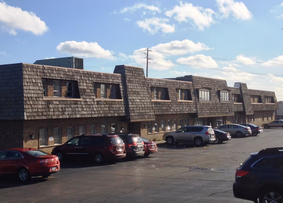 Exterior view of a multi-unit senior living facility with a long, shingled roof and parked cars.