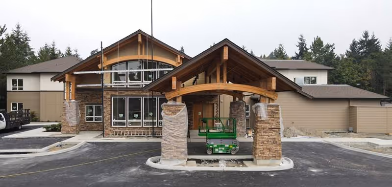 Front exterior view of Silverado Bellevue Memory Care Community building under construction with a covered entrance supported by stone pillars and wooden beams, surrounded by newly paved parking area and trees in the background.