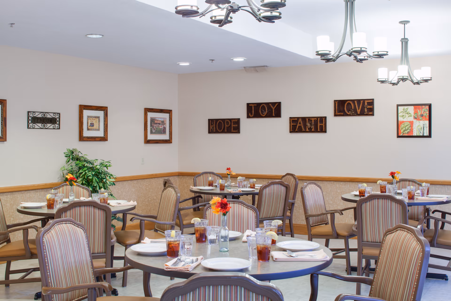 A dining room with round tables set with plates, glasses of iced tea, napkins, and silverware. The chairs have striped upholstery and wooden armrests. The walls are decorated with framed pictures and wooden plaques with the words HOPE, JOY, FAITH, and LOVE. There are ceiling lights and a potted plant in the corner.