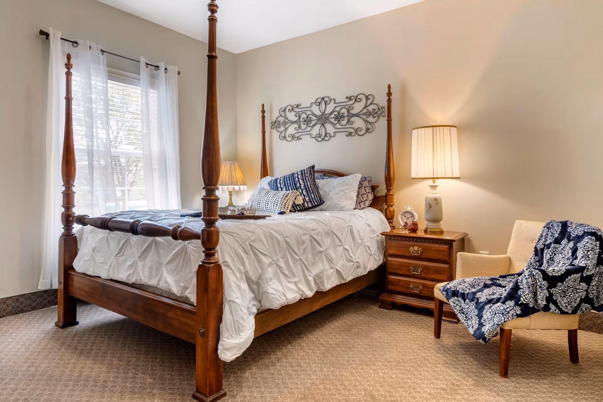 A cozy bedroom with a wooden four-poster bed dressed in white bedding and multiple decorative pillows. Next to the bed is a wooden nightstand with a lamp, an alarm clock, and a small decorative item. A beige armchair with a blue and white patterned throw blanket is positioned nearby. Light filters in through a window with sheer white curtains, and a decorative metal wall art piece hangs above the bed.
