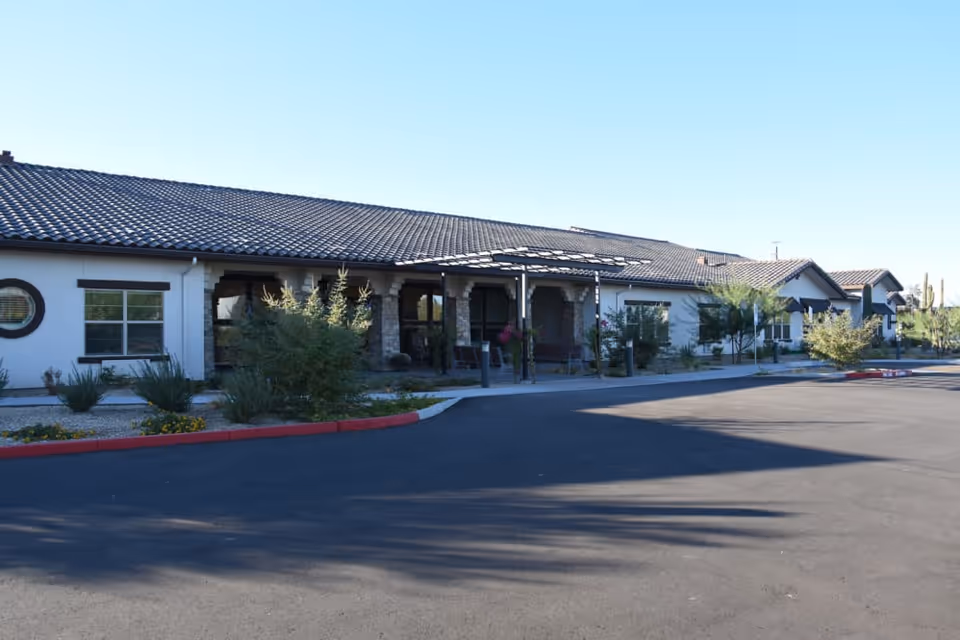 Exterior view of a single-story building with a tiled roof and a covered entrance supported by stone columns. There are windows and desert landscaping with bushes and small trees in front of the building, along with a paved driveway and parking area under a clear blue sky.