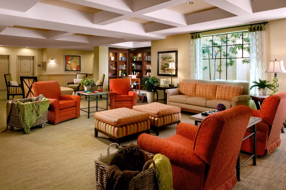 A cozy living room area with orange armchairs, a beige sofa with orange cushions, two striped ottomans, side tables with lamps, a large window with sheer curtains, and a bookshelf filled with books in the background.