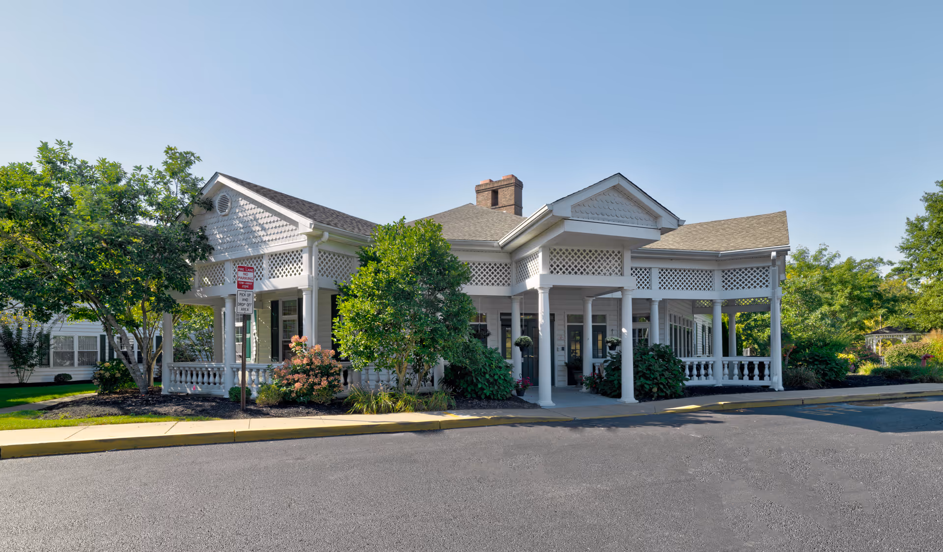 Entrance of a white single-story senior living building with a covered porch, columns, and landscaped shrubs.