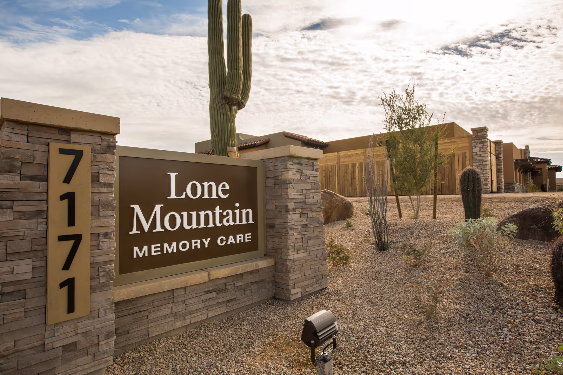 Stone entrance sign reading 'Lone Mountain Memory Care' beside address numbers with desert landscaping, cacti, and the facility building in the background.