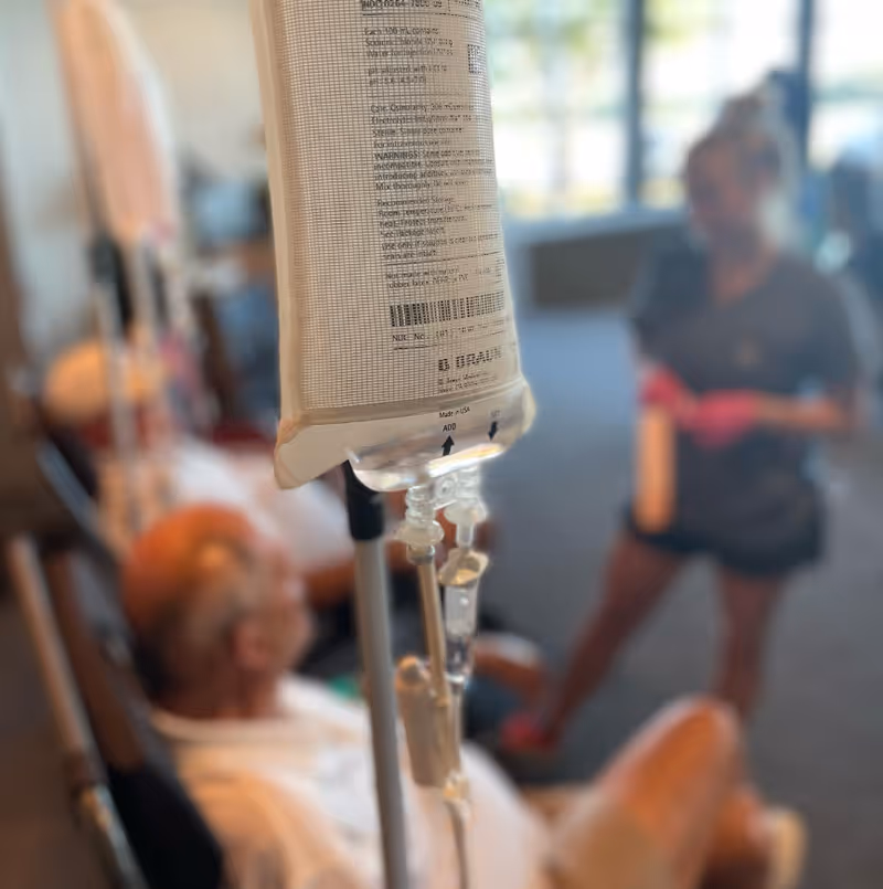 An IV drip bag hangs in focus in a care room with a resident.