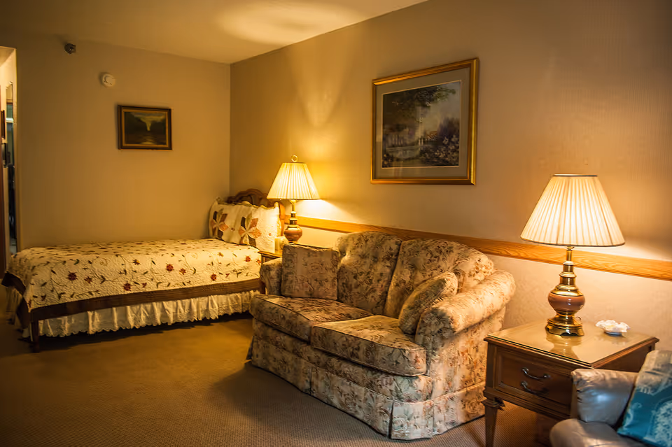 A cozy senior living room with a floral patterned loveseat, a wooden side table with a lamp, and a single bed with a floral quilt. The walls are decorated with framed paintings, and the room is warmly lit by two lamps.