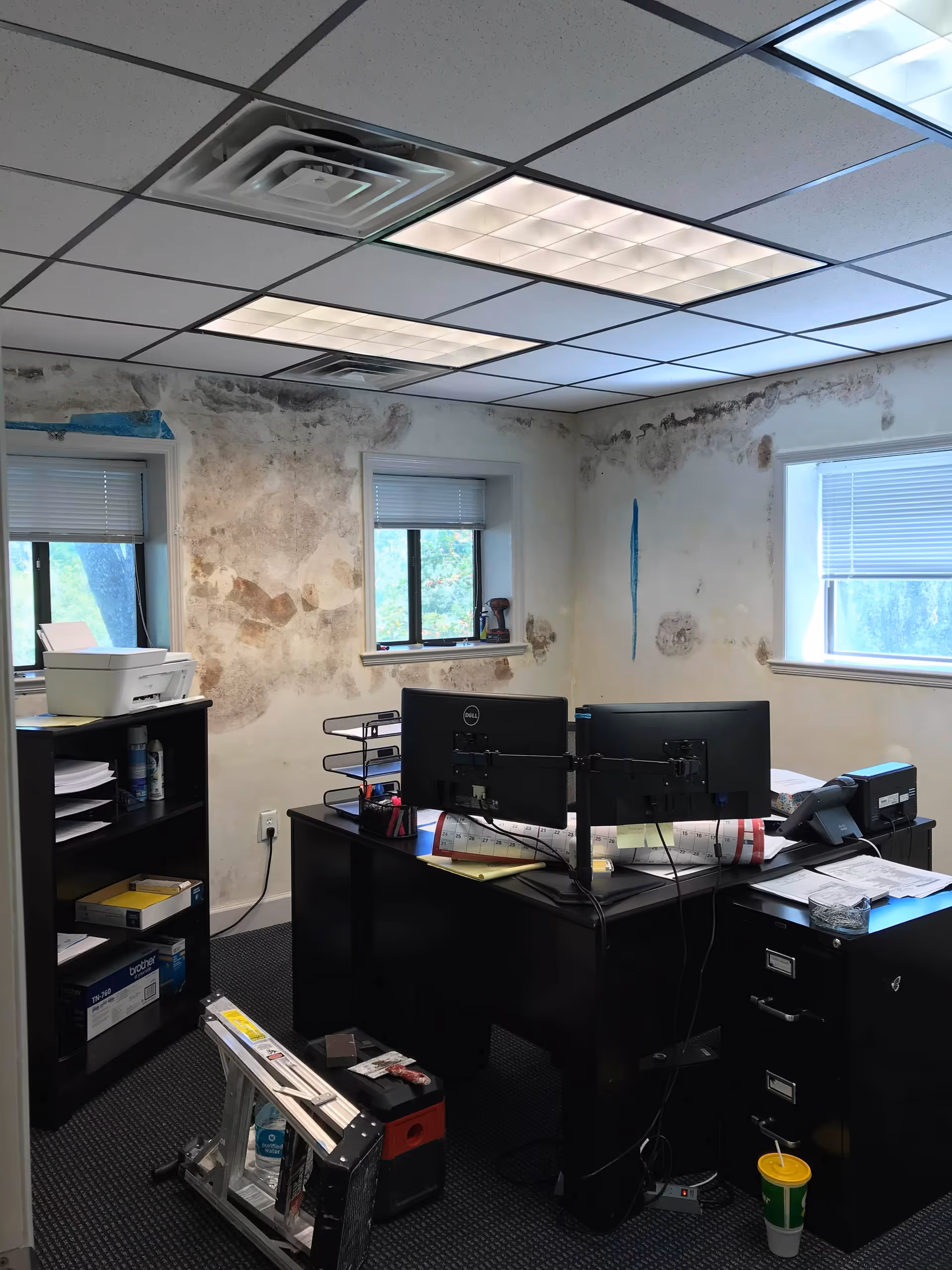 Office desk with two monitors and scattered papers, alongside visible damaged plaster walls.