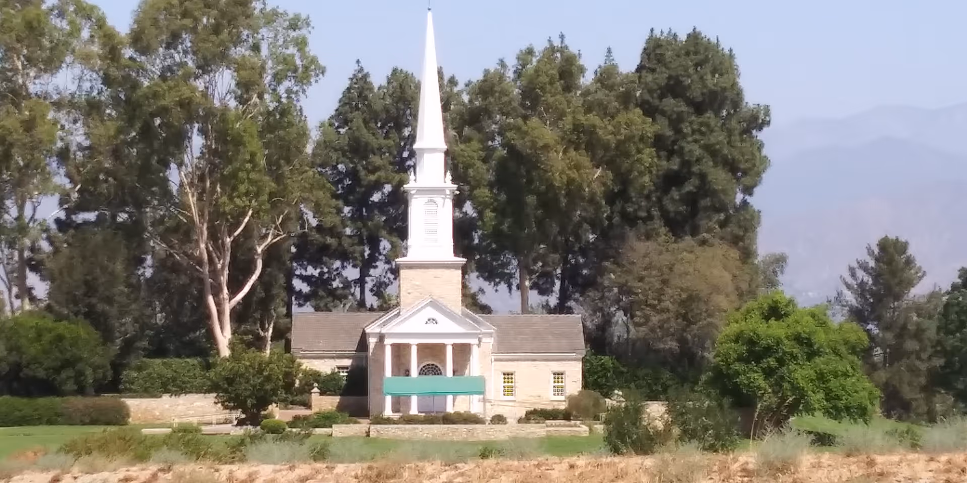 Exterior view of a small church-like building with a tall steeple, surrounded by trees.