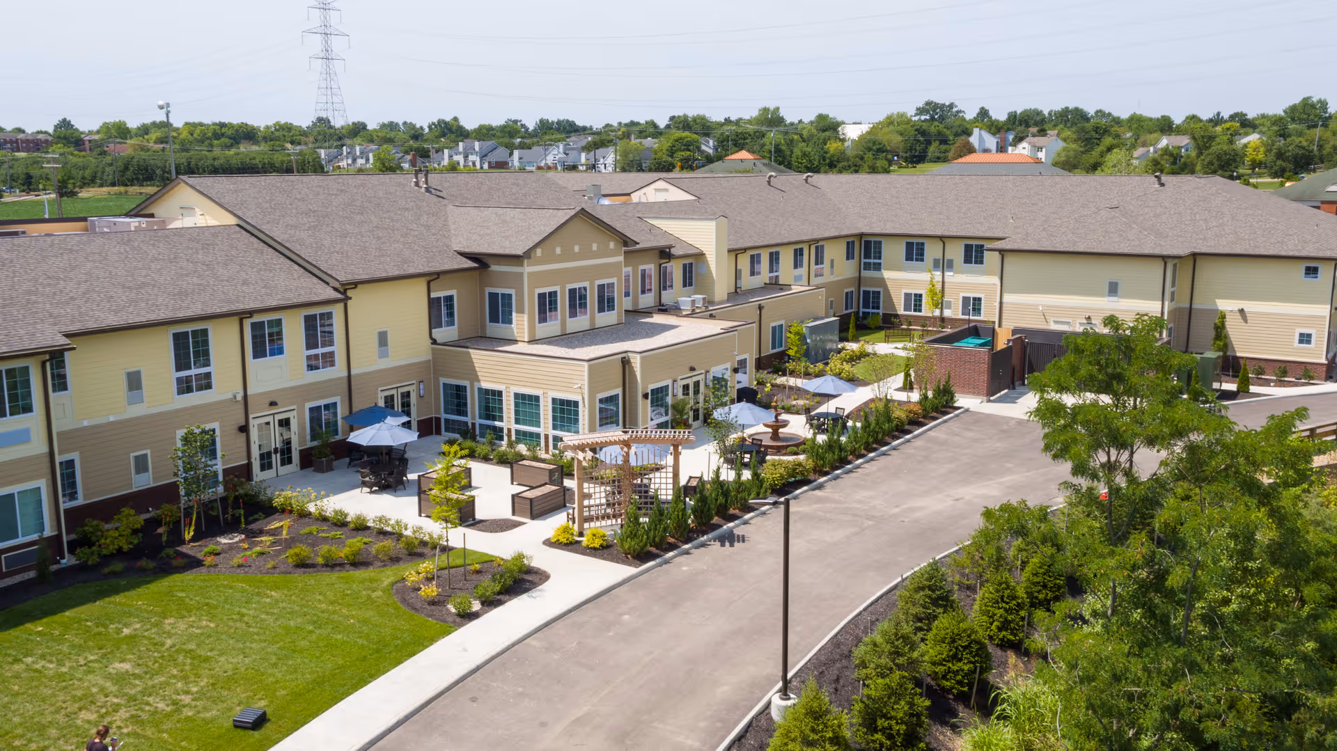 Aerial view of Mason Assisted Living & Memory Care facility showing a large two-story building with beige and yellow exterior walls, multiple windows, and a brown roof. The facility features outdoor seating areas with umbrellas, landscaped gardens, and a driveway surrounded by greenery and trees.