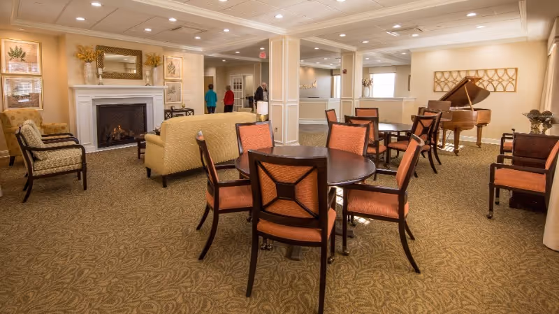Well-lit senior living community lounge with round tables and chairs, a fireplace, sofa seating, and a grand piano.