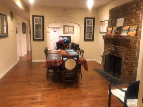 A dining room with a table and chairs, brick fireplace, and resident at the far end.