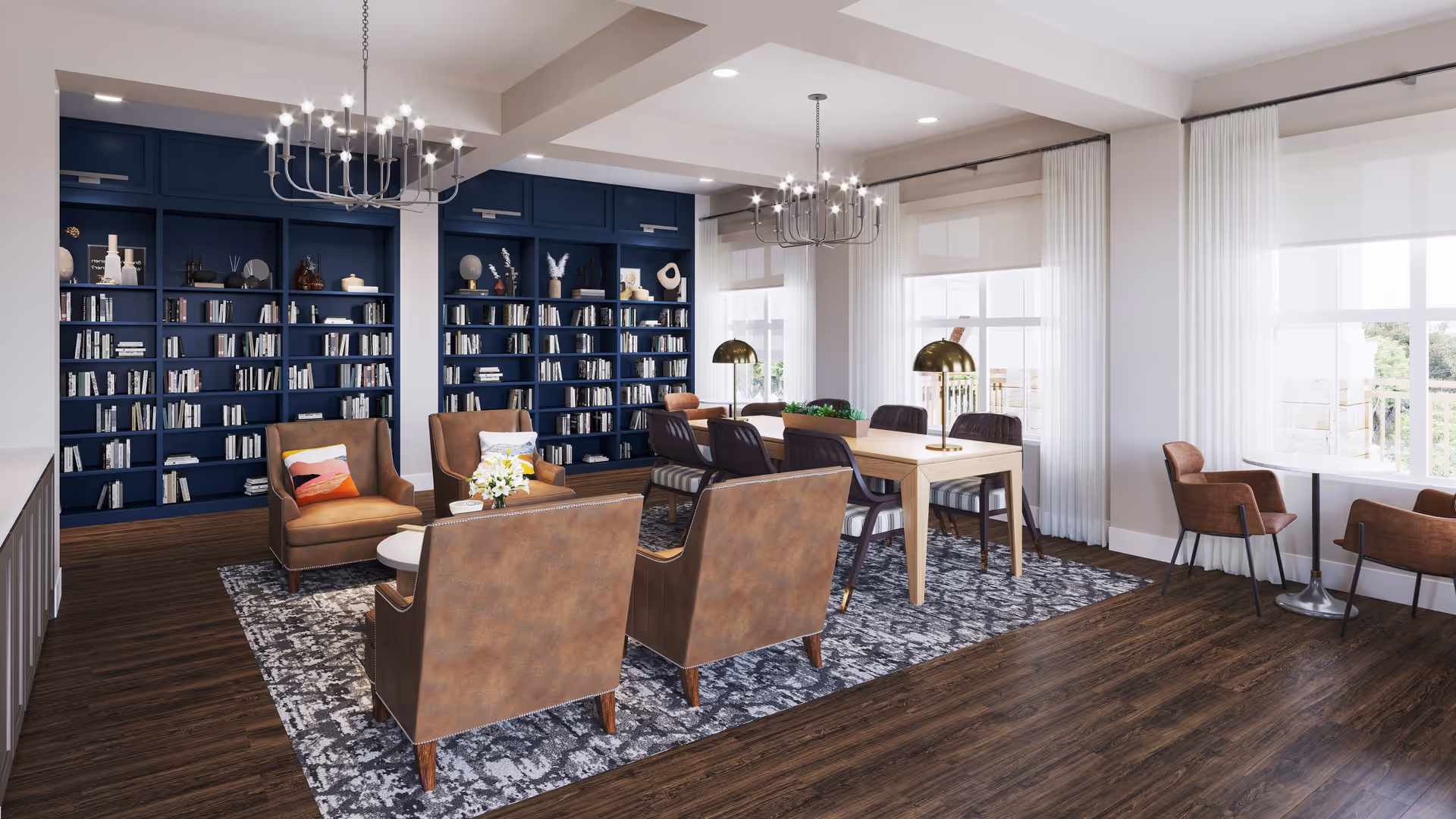A bright and spacious common area with dark wood flooring, featuring a seating arrangement of four brown leather armchairs around a small round table with a flower vase. Behind the seating area is a large blue bookshelf filled with books and decorative items. To the right, there is a wooden table with six chairs and two brass table lamps. Large windows with white curtains allow natural light to fill the room.