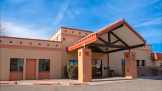 Entrance of a single-story stucco building with a covered porte-cochere, red-tile accents, and a clear blue sky.