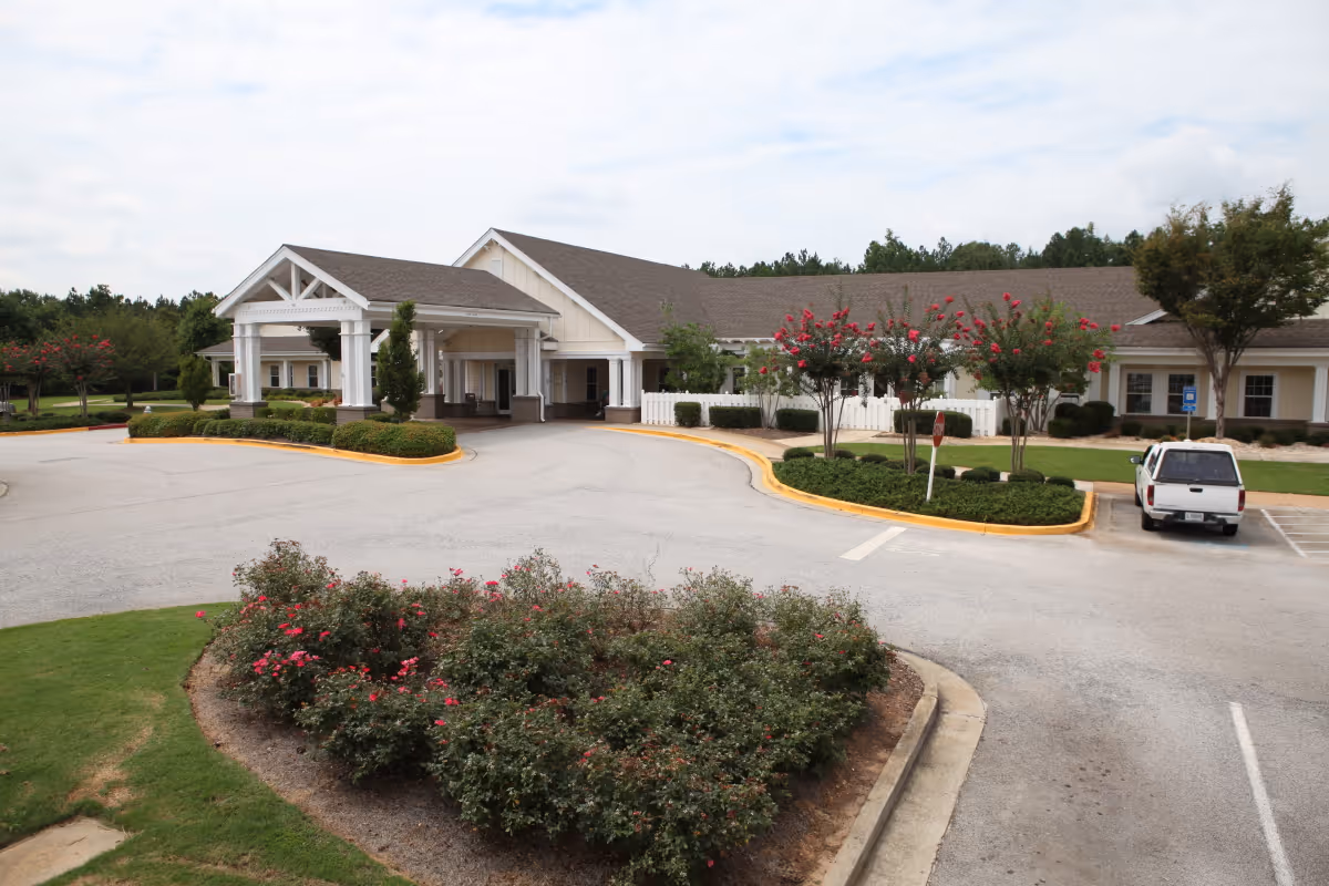 Front entrance of a single-story senior living facility with a covered porte-cochere, circular driveway, landscaped islands with flowering shrubs, and a parked white pickup truck.