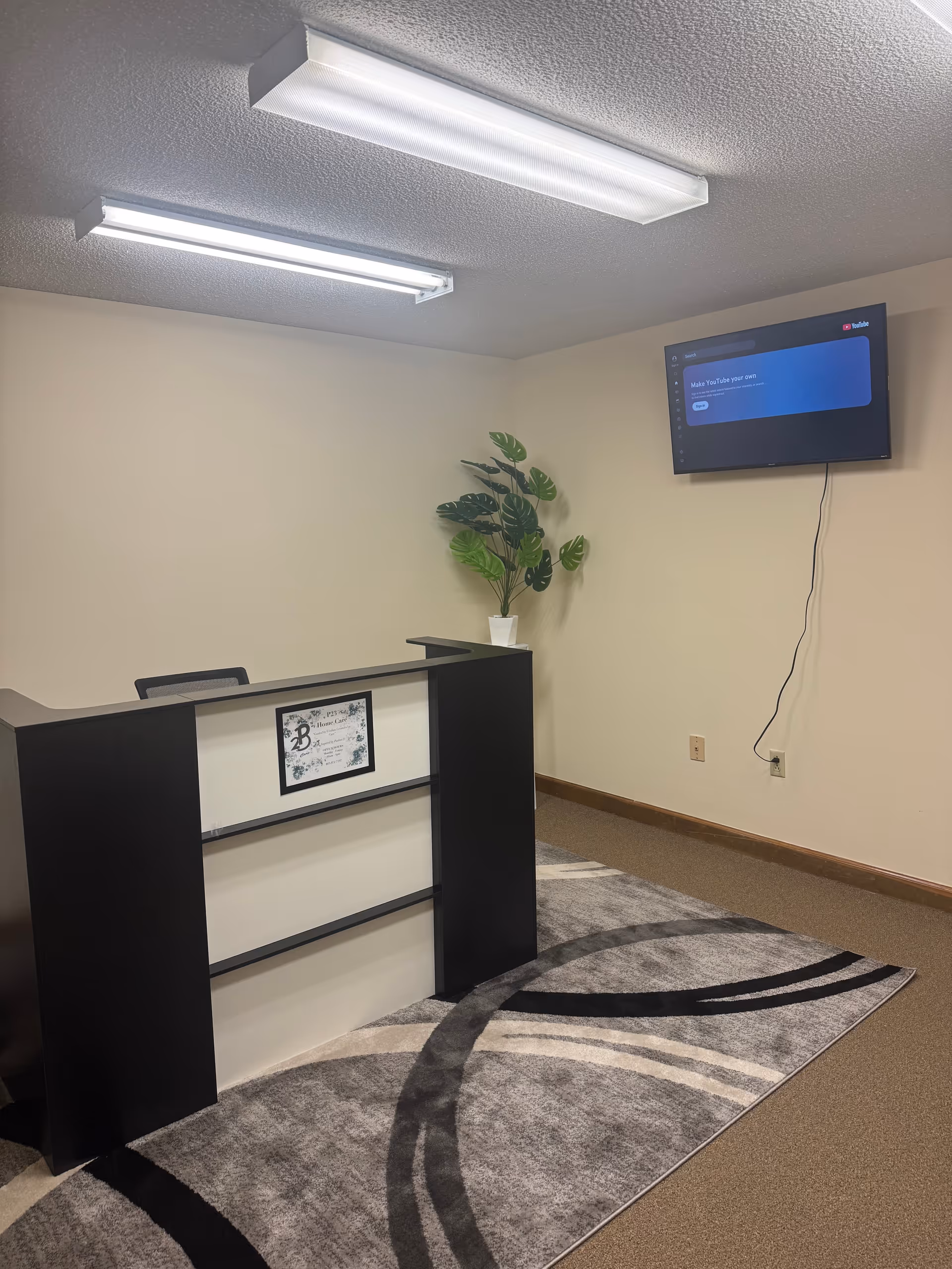 Reception desk in a neutral lobby with plant, rug, and wall-mounted TV.