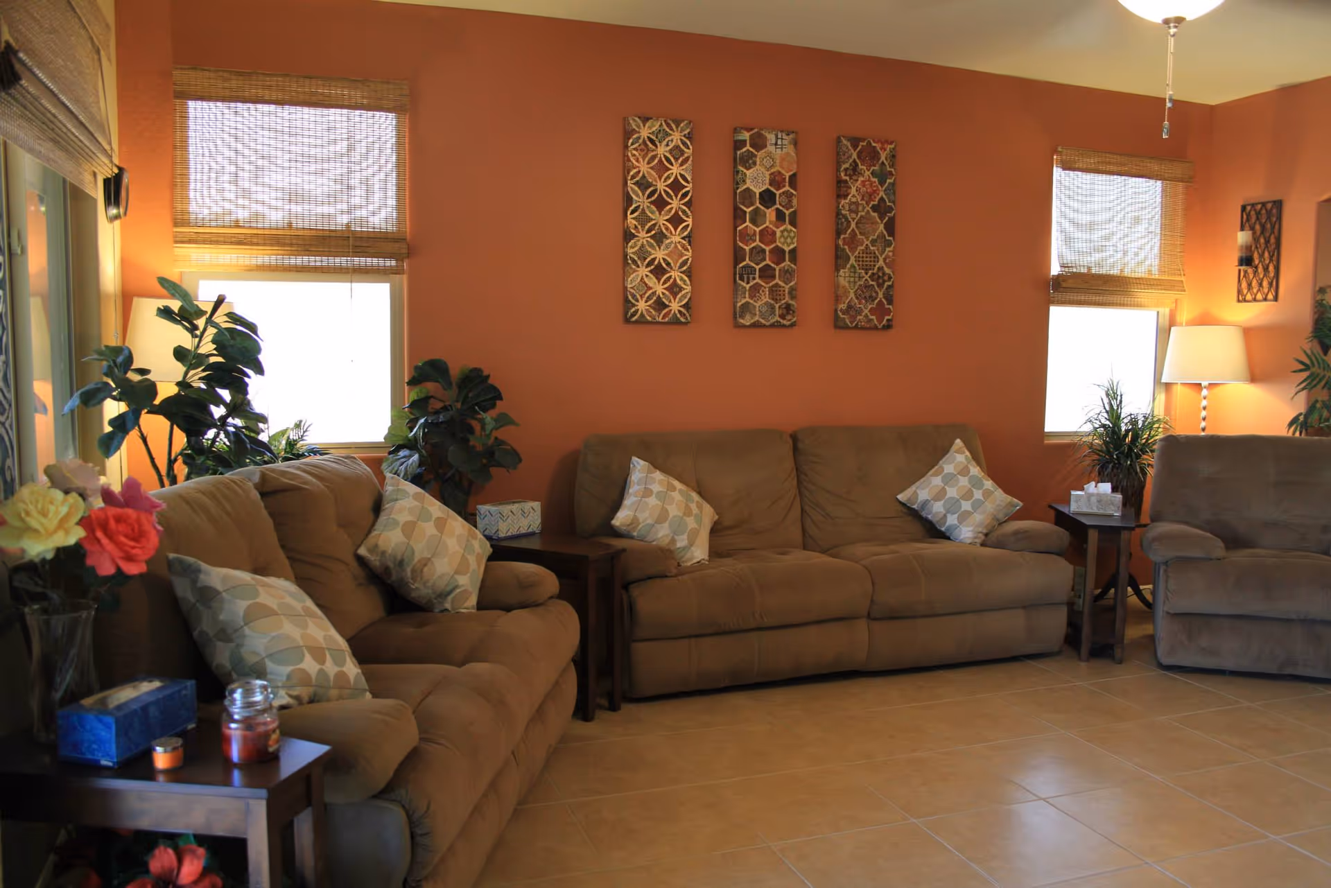 A cozy living room with three brown sofas adorned with patterned cushions. The walls are painted orange and decorated with three vertical patterned wall hangings. There are two windows with bamboo blinds, several green plants, a floor lamp, and side tables holding a tissue box, candles, and flowers.