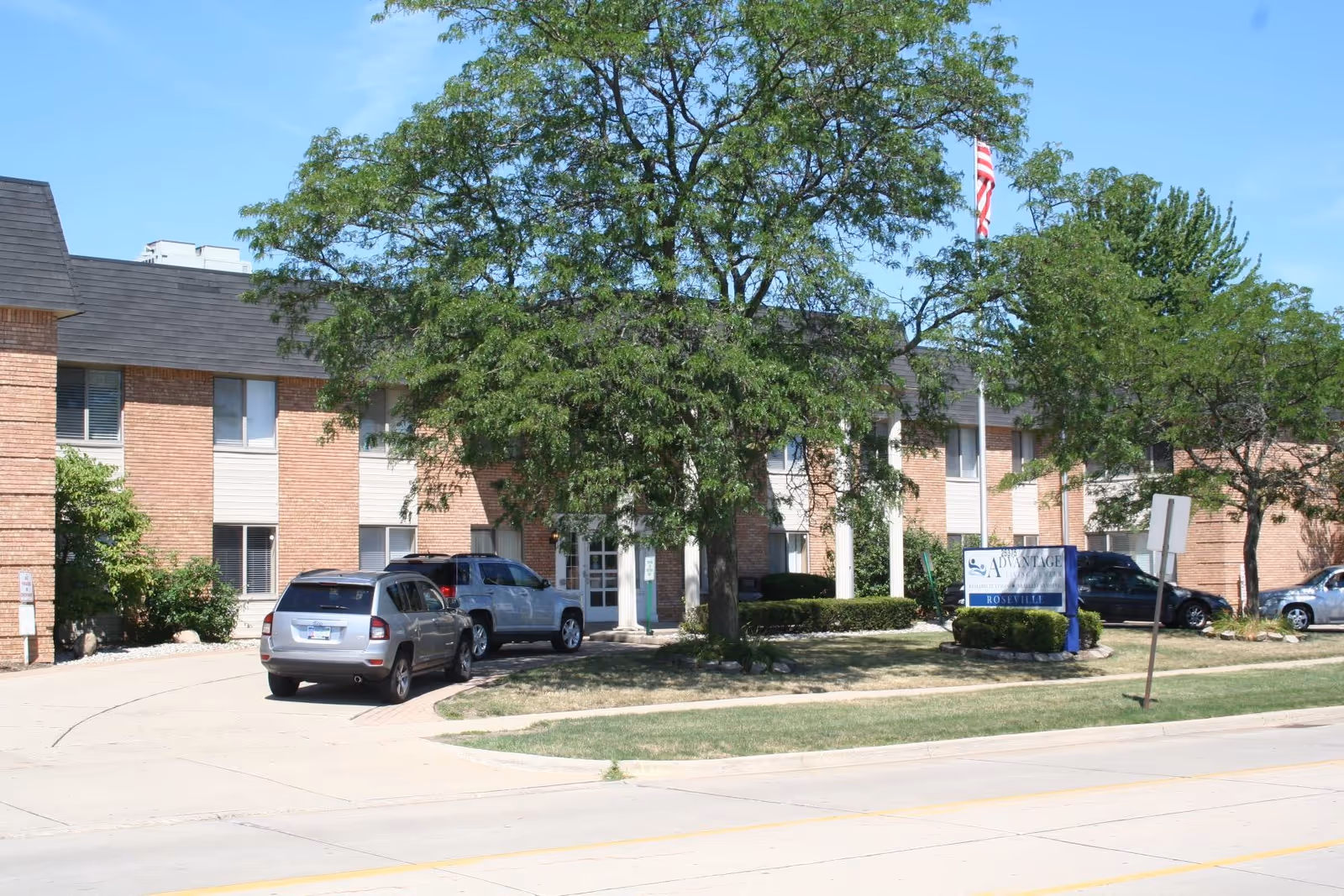 Front view of a two-story brick senior living building with cars in the driveway, trees, and a roadside sign.