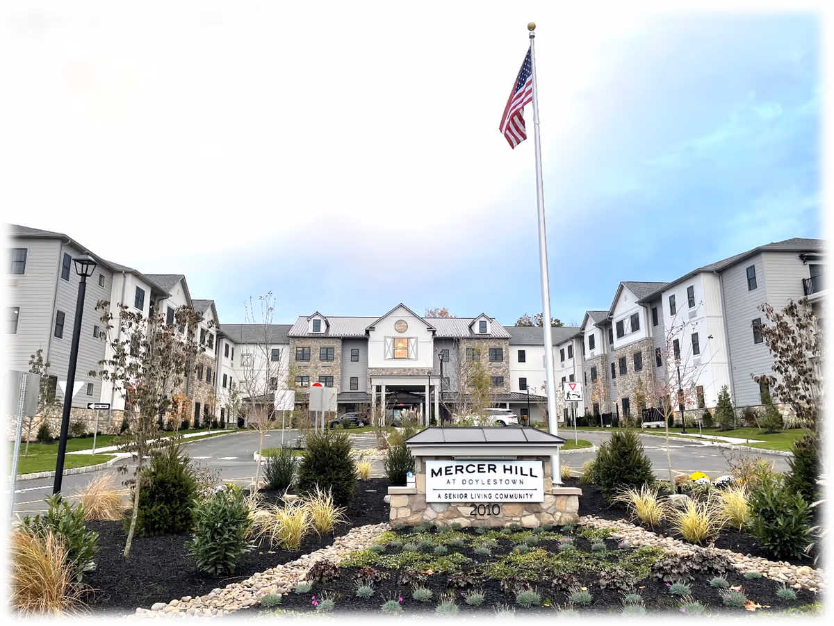 Front exterior of the Mercer Hill at Doylestown senior living building with landscaped entrance and an American flag on a tall flagpole.